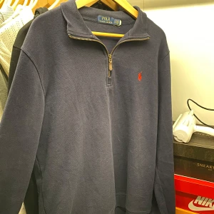 Mörkblå tröja från Polo Ralph Lauren - Säljer en snygg mörkblå tröja från Polo Ralph Lauren med dragkedja vid halsen. Den har en klassisk design med det ikoniska röda logotypen på bröstet. Perfekt för både vardag och lite finare tillfällen. Passar bra till jeans eller chinos!