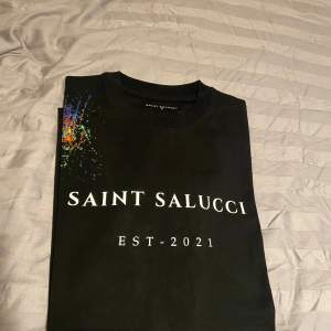 Snygg svart t-shirt från Saint Salucci med tryck 'EST. 2021' på framsidan. Den har en cool färgexplosion på axeln och en gul detalj med knapp längst ner. Perfekt för en casual look! Storlek S.M. L . XL 