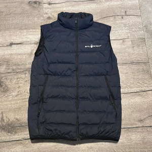 Mörkblå dunväst från Sail Racing - Säljer en mörkblå dunväst från Sail Racing i mycket bra skick. Västen har en dragkedja framtill och är fylld med högkvalitativt dun från Allied Feather + Down. Perfekt för kyliga dagar och har en stilren design med logga på bröstet och ryggen.