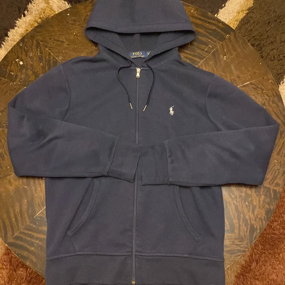 Säljer en snygg mörkblå hoodie från Polo Ralph Lauren i mycket bra skick. Den har en dragkedja framtill och den klassiska loggan broderad på bröstet. Perfekt för höst och vinter, med en skön passform och långa ärmar. Huvan ger extra mysfaktor! Ny pris 1500kr. Tröjor & Koftor.