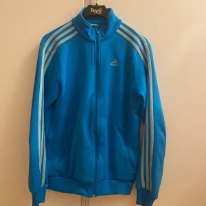 Blå träningsjacka från Adidas - Säljer en snygg blå träningsjacka från Adidas i mycket bra skick. Jackan har de klassiska tre ränderna längs ärmarna och en dragkedja framtill. Perfekt för träning eller som en casual vardagslook. Den är långärmad och gjord i ett bekvämt syntetmaterial.