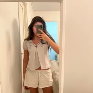  blus med volangdetaljer - Säljer en söt blus med korta ärmar och volangdetaljer vid halsen. Blusen har en liten knytning framtill och är perfekt för sommardagar. Den är i ett lätt och luftigt material, vilket gör den bekväm att bära.