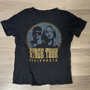 Svart t-shirt med tryck från Gina Tricot - Cool svart t-shirt från Gina Tricot i storlek XS. Den har ett tryck med texten 'Stage Tour California' och ett motiv av två personer. Perfekt för en avslappnad stil eller konsertkvällar. T-shirten är kortärmad och i bra skick.