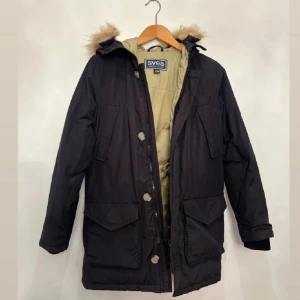 Svart parkajacka från Svea - Säljer en svart parkajacka från Svea i mycket bra skick. Jackan har en mysig pälsfodrad huva och stängs med både dragkedja och knappar. Perfekt för kalla vinterdagar med sina långa ärmar och praktiska fickor. En klassisk och stilren design som passar till det mesta.