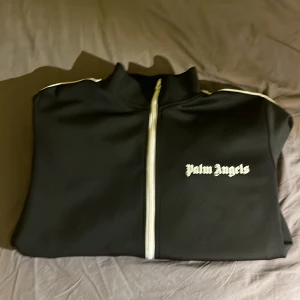 Palm Angels zip - Säljer en snygg svart zip från Palm Angels. Den har vita ränder längs ärmarna och en dragkedja framtill. Perfekt för en casual look. Zipen är i mycket bra skick.