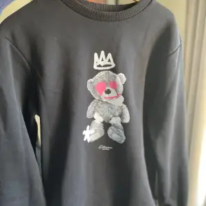 Snygg svart sweatshirt med tryck av en teddybjörn med krona och hjärtformade glasögon. 