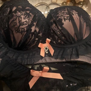 Svart spets-bh med rosett - Säljer en elegant svart bh med vacker spets och broderade detaljer. Den har en söt rosa rosett med en liten pärla framtill. Bh:n är vadderad och har justerbara band för perfekt passform. Perfekt för speciella tillfällen eller när du vill känna dig extra fin.