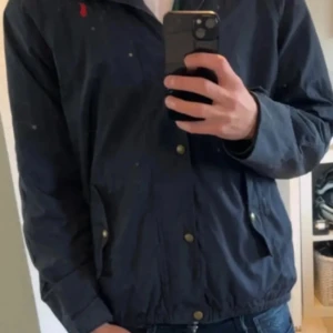 Svart jacka från Ralph Lauren - Säljer en snygg svart jacka från Ralph Lauren i storlek XL i barnstorlek, skulle säga att den ligger vid S-M i herr storlek. Jackan har en klassisk design med broderad röd logga på bröstet och knappar framtill. Perfekt för höst och vår, med en stilren look som funkar till det mesta. Passar både till vardags och lite finare tillfällen.