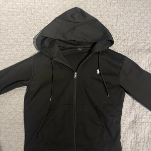 Svart polo zip hoodie - Säljer en svart huvtröja från Polo Ralph Lauren i mycket bra skick. Den har en dragkedja framtill och en liten broderad logga på bröstet. Perfekt för höst och vår, med en skön passform och långa ärmar. Huvan har dragsko för justering.