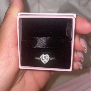 Pandoraring med hjärtformad sten (äkta) - Säljer en vacker pandoraring med en glittrande hjärtformad sten i mitten. Ringen har små stenar längs bandet som ger extra bling. Perfekt för speciella tillfällen eller som en kärleksgåva. Kommer i en söt rosa ask. 💍✨använd ett par gånger. Storlek 48. Nypris 899kr! 