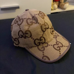 Beige keps från Gucci - Snygg beige keps från Gucci med det ikoniska GG-mönstret i brunt. Kepsen har en klassisk design med böjd skärm och justerbar passform. Perfekt för att ge din outfit en lyxig touch.