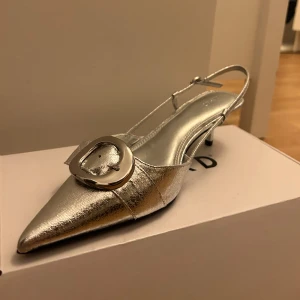 Silvriga pumps med spänne - Eleganta silvriga pumps från NA-KD med spetsig tå och dekorativt spänne. Perfekta för en festlig kväll. Skorna har en smal klack och en justerbar rem runt hälen.