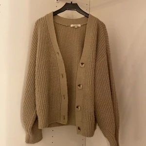 Beige kofta från Samsa Young - Mysig beige kofta från Samsa Young med stora knappar framtill. Den har en ribbad struktur och långa ärmar, perfekt för kyligare dagar. Koftan är både stilren och bekväm, ett måste i garderoben!