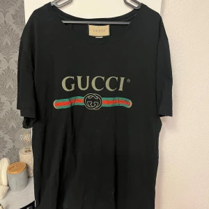 Svart t-shirt från Gucci - Snygg svart t-shirt från Gucci med det ikoniska logotyptrycket i vitt, rött och grönt på framsidan. Perfekt för en stilren och lyxig look. 