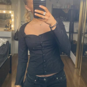 Svart långärmad topp med knappar - Snygg svart långärmad topp med knappar framtill. Toppen har en tight passform och en stilren design som passar perfekt till både vardag och fest. Perfekt för höst och vinter!