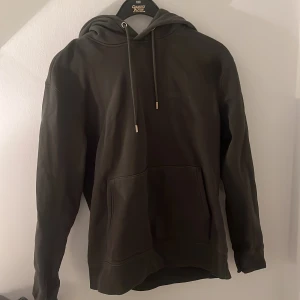 J Lindeberg Mörkgrön hoodie - Säljer en stilren mörkgrön hoodie med justerbar huva och stor magficka. Perfekt för en avslappnad look. Storlek S