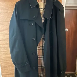 Svart trenchcoat från Burberry - Säljer en elegant svart trenchcoat från Burberry, modell The Kensington. Jackan har ett klassiskt rutigt foder och knappar framtill. Perfekt för en stilren look. Använd 1 gång så i nyskick.