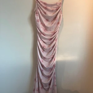 Rosa blommig långklänning - Elegant rosa långklänning med blommigt mönster och smala axelband. Klänningen har en draperad design som ger en vacker siluett.