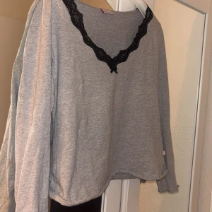 Grå tröja med spetsdetalj från Miss Triumph - Säljer en grå tröja från Miss Triumph med svart spetsdetalj vid halsringningen. Fick den av en vän så den har nog använts många gånger och slutet av ärmen och vid änden av tröjan viker den upp sig lite, ser då ut att vara klippt sen tidigare.