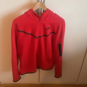 Röd hoodie från Nike - Säljer en snygg röd hoodie från Nike med dragkedja och huva. Den har svarta detaljer och är perfekt för en sportig look. Passar bra för kyligare dagar.Den är i barnstorlek XL