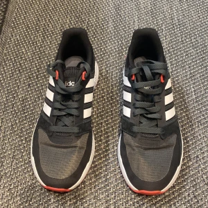 Svarta Adidas sneakers, 39  - Snygga svarta Adidas sneakers med klassiska vita ränder och röda detaljer. Skorna har snörning och en bekväm sula, perfekt för vardagsbruk.