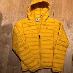 Gul dunjacka från Hang Ten - Säljer en snygg gul dunjacka från Hang Ten med huva och dragkedja. Perfekt för kyliga dagar med sin quiltade design. Jackan har en normal passform och är lätt att bära.