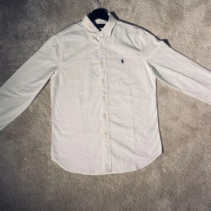 Vit skjorta från Ralph Lauren  - Säljer en klassisk vit skjorta från Ralph Lauren i storlek S. Skjortan är i bomull och har långa ärmar med knappar framtill. Den har en broderad logga i blått på bröstet. Perfekt för både vardag och fest! 