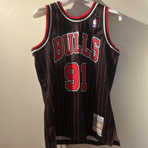 Svart och röd basketlinne från Mitchell & Ness - Säljer en svart och röd basketlinne från Mitchell & Ness med 'Bulls' och nummer 91 på framsidan. Baksidan har 'Rodman' och nummer 91. Linnet är en del av Hardwood Classics-kollektionen och har ett randigt mönster.