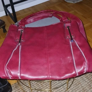 Röd axelväska med dragkedjor, innerdragkedja. - Snygg röd axelväska i skinn med stilrena dragkedjedetaljer. Perfekt för att ge en färgklick till din outfit. Väskan har två handtag och en praktisk storlek för vardagsbruk.