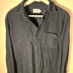 Svart overshirt från East West - Snygg svart overshirt från East West med knappar framtill och en bröstficka. Perfekt för lager-på-lager-stil. Passar bra till både vardag och mer avslappnade tillfällen.