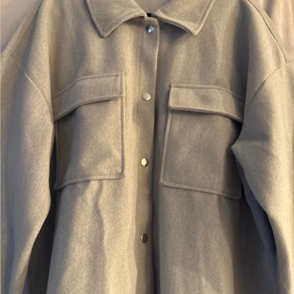 Snygg grå overshirt från Gina Tricot med knappar framtill och två stora bröstfickor. Perfekt för lager-på-lager-stil. Den har en klassisk krage och långa ärmar.. Takit.