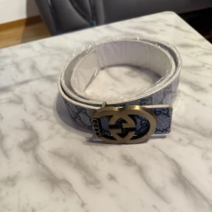 Mönstrat bälte med metallspänne - Snyggt bälte med ett distinkt mönster i blått och vitt. Bältet har ett stort metallspänne i guldton. Perfekt för att ge en stilren touch till din outfit.