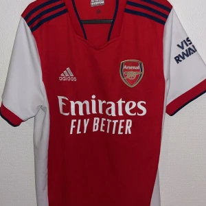 Röd Arsenal fotbollströja från Adidas - Snygg röd Arsenal fotbollströja från Adidas med vita ärmar och blå detaljer. Tröjan har klubbens emblem på bröstet och sponsorlogga framtill. Perfekt för alla Arsenal-fans!
