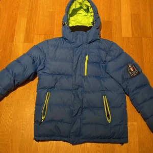 Blå dunjacka från Jean Paul - Säljer en blå dunjacka från Jean Paul med huva och dragkedjor i kontrasterande gul färg. Jackan har en broderad logga på ärmen och flera praktiska fickor med dragkedja. Perfekt för kyliga dagar.