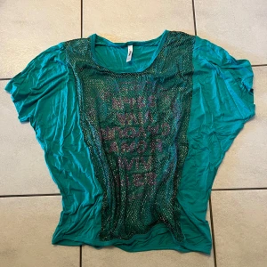 Turkos topp med nät och text - Säljer en turkos topp med ett nätlager framtill och text i rosa. Toppen har en lös passform och korta ärmar. Perfekt för en avslappnad stil.