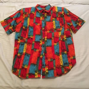 Färgglad kortärmad skjorta - Säljer en färgglad kortärmad skjorta med ett unikt mönster i rött, blått, gult och svart. Skjortan har knappar framtill och en bröstficka. Perfekt för att sticka ut i mängden!