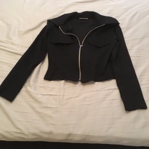 Svart croppad jacka med dragkedja - Snygg svart croppad jacka med dragkedja framtill och långa ärmar. Perfekt för en trendig look.