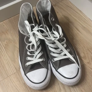 Bruna sneakers från Converse - Säljer ett par klassiska bruna Converse sneakers med vita snören och gummisula. Skorna har en hög modell med den ikoniska loggan på sidan. Perfekta för en avslappnad stil. Orginalpris 480kr