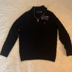 Svart tröja med dragkedja från Ralph Lauren - Säljer en stilren svart tröja från Ralph Lauren med dragkedja och röd logotyp på bröstet. Tröjan har långa ärmar och en ribbad struktur som ger en klassisk look. Perfekt för kyligare dagar. Tröjan är storlek L men passar M. Nypris 2500 mitt pris 500