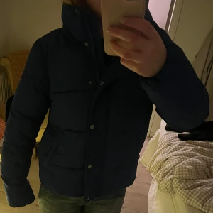 Mörkblå dunjacka - Säljer en mörkblå dunjacka med dragkedja och knappar. Jackan har en pufferdesign som ger extra värme och stil. Perfekt för kyliga dagar.
