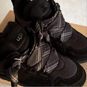 Svarta sneakers från UGG - Snygga svarta sneakers från UGG med robust design och breda snören. Skorna har en modern look med en kombination av olika material och en bekväm sula. Slutsålda överallt, kartong och extra svarta snören följer med. 