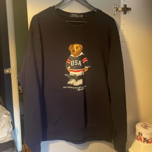 Polo bear sweatshirt  - Säljer en svart långärmad tröja från Polo Ralph Lauren med ett coolt björntryck som bär en USA-tröja. Tröjan har en klassisk passform och är tillverkad i mjukt material.