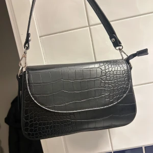 Svart axelväska från NA-KD - Snygg svart axelväska i krokodilpräglat mönster från NA-KD. Väskan har en justerbar axelrem och en stilren design med lock. Perfekt för att ge en trendig touch till din outfit.