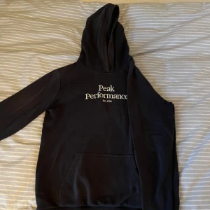 Svart hoodie från Peak Performance - Säljer en svart hoodie från Peak Performance med vit text på bröstet. Den har en klassisk känguruficka och en bekväm huva. Lite solblekt också så syns att den är använd. Storlek 170 S men passar också XS. 