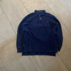 Blå rugbytröja från Ralph Lauren - Säljer en klassisk blå Rugbytröja från Ralph Lauren med lång ärm och den ikoniska röda loggan på bröstet. Perfekt för en stilren look. Tröjan har en krage med knappar och är i storlek XS men passar S
