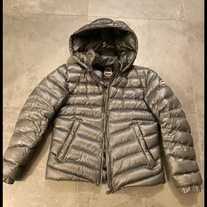 Grå dunjacka från Colmar - Snygg och varm grå dunjacka från Colmar med huva och dragkedja. Perfekt för kyliga dagar. Jackan har en quiltad design och långa ärmar. Nypris 6500kr