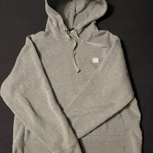 Acne Studios hoodie (grå) - Säljer en stilren grå hoodie med dragkedja och justerbar huva. Perfekt för en avslappnad look. Den har en liten fyrkantig logga på bröstet och en stor ficka framtill.