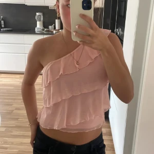 Rosa oneshoulder topp - Säljer en söt rosa oneshoulder topp med volanger. Perfekt för sommarens varma dagar. Toppen har en luftig passform och ärmlös design.