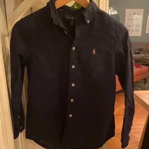 Mörkblå skjorta från Ralph Lauren - Snygg mörkblå skjorta från Ralph Lauren med klassisk krage och knappar framtill. Skjortan har en broderad logotyp på bröstet och är perfekt för en stilren look. !skjortan är en barnstorlek och passar för dem som är runt 150cm!