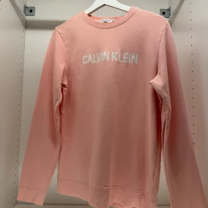Rosa sweatshirt från Calvin Klein - Säljer en snygg rosa sweatshirt från Calvin Klein med deras logga tryckt på framsidan. Tröjan har långa ärmar och en rund halsringning. Perfekt för en avslappnad stil.